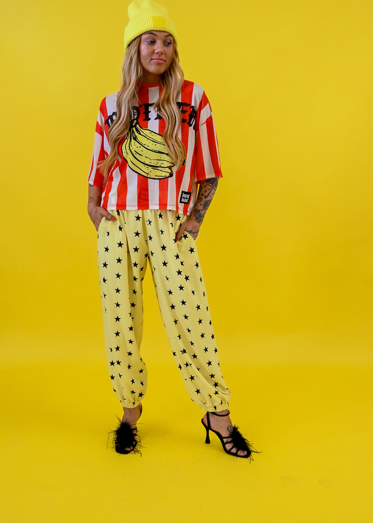 A model stands against a solid bright yellow studio background wearing a yellow beanie, a red and white striped graphic mesh jersey with a banana print, yellow jogger pants with black star patterns, and black strappy heels