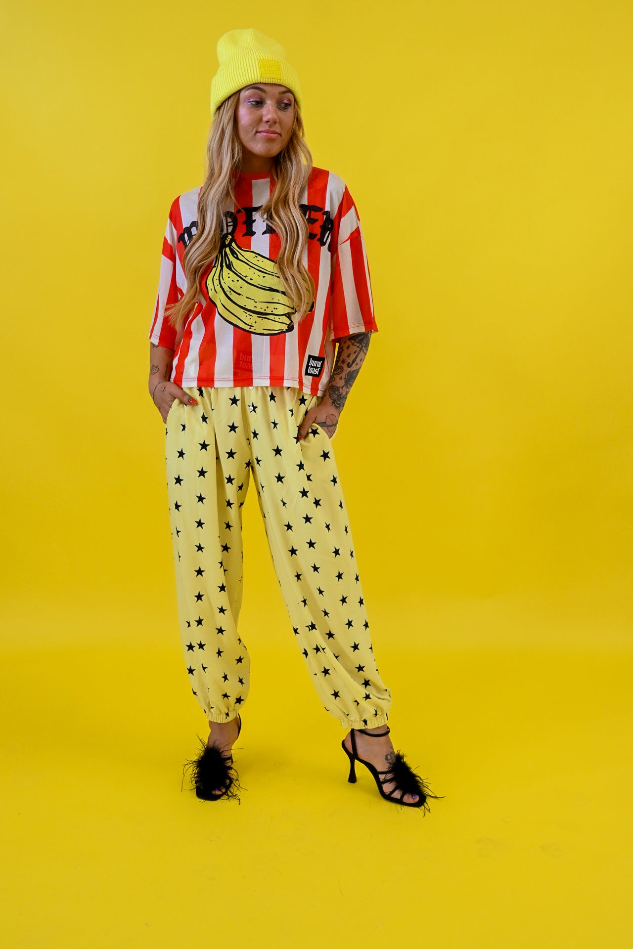 A model stands against a solid bright yellow studio background wearing a yellow beanie, a red and white striped graphic mesh jersey with a banana print, yellow jogger pants with black star patterns, and black strappy heels
