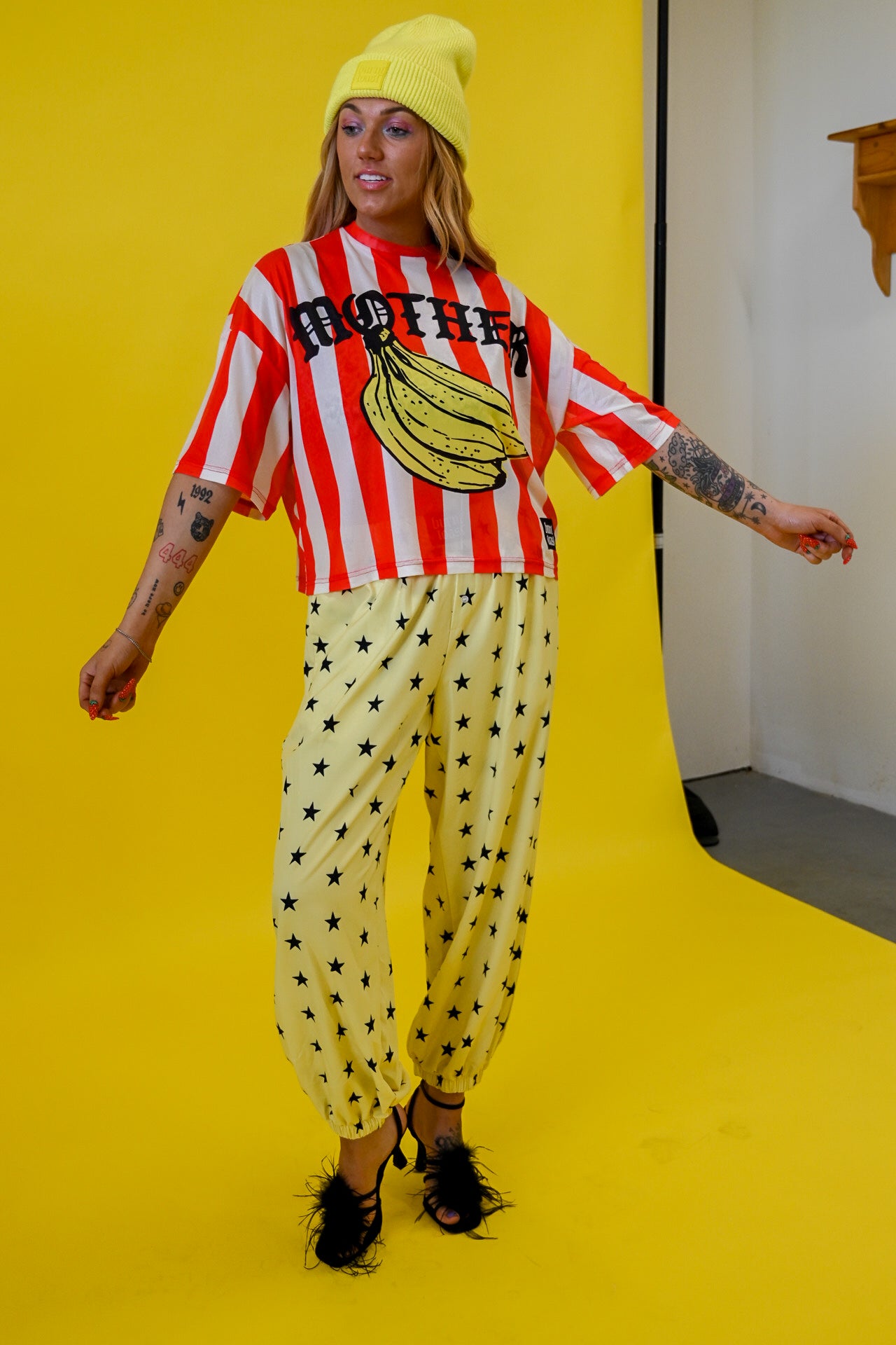 A model wearing a retro outfit featuring a red and white striped mesh jersey with a banana graphic and the word 'MOTHER', paired with yellow pants patterned with black stars, a yellow beanie, and black fuzzy shoes, set against a bright yellow studio backdrop
