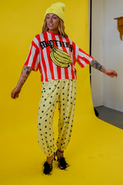 A model wearing a retro outfit featuring a red and white striped mesh jersey with a banana graphic and the word 'MOTHER', paired with yellow pants patterned with black stars, a yellow beanie, and black fuzzy shoes, set against a bright yellow studio backdrop