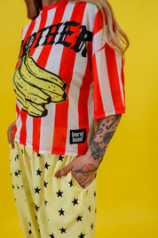 A person wearing a red and white vertically striped graphic T-shirt with a large yellow banana illustration and the word 'MOTHER' across the chest, paired with pale yellow pants covered in black stars. The photo is taken against a solid bright yellow background