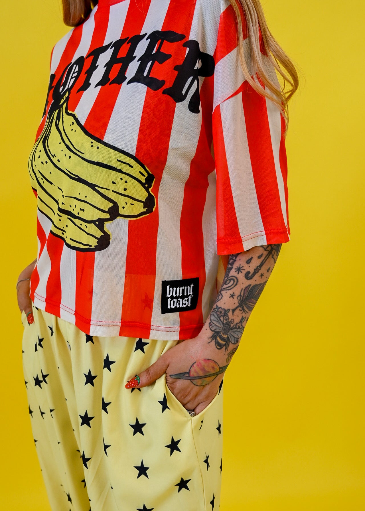 A person wearing a red and white vertically striped graphic jersey with a large yellow banana illustration and the word 'MOTHER' across the chest, paired with pale yellow pants covered in black stars. The photo is taken against a solid bright yellow background