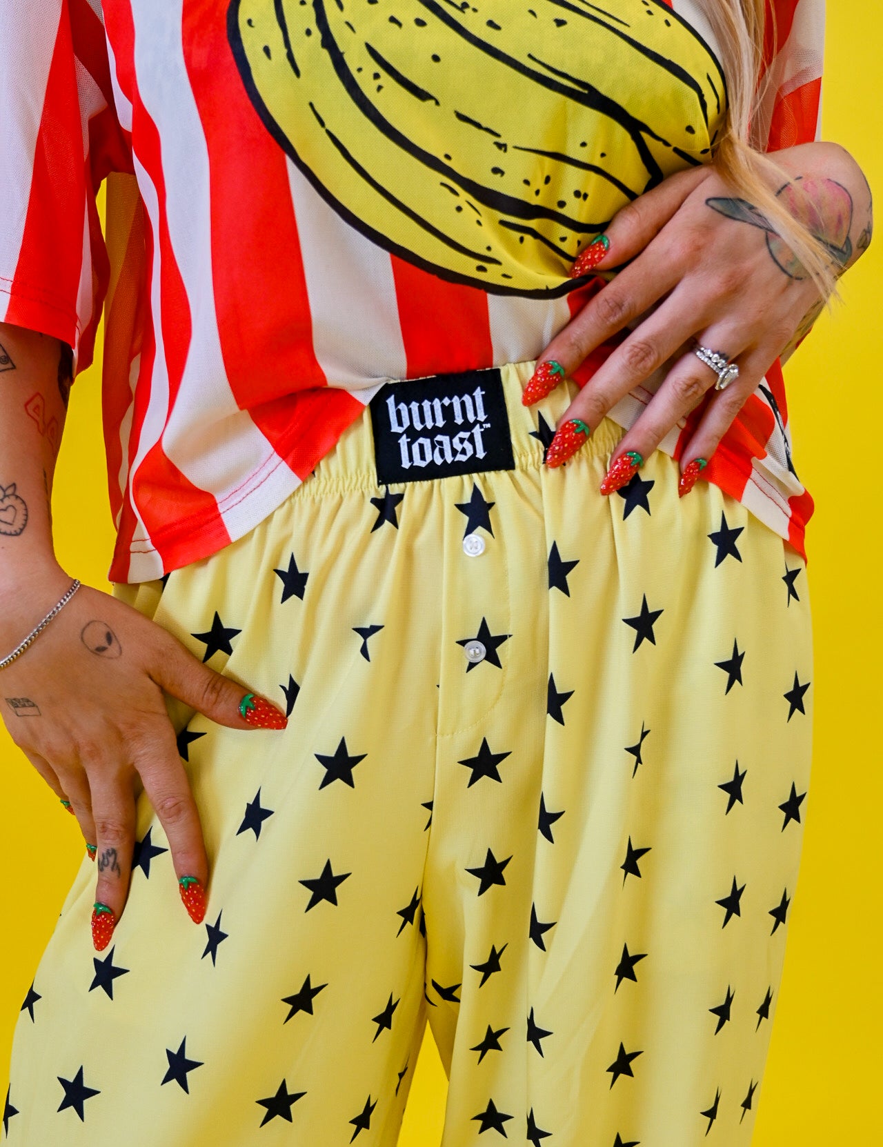 A person wearing yellow pants with a black star pattern and a red-and-white striped shirt with a banana graphic, standing against a bright yellow background. A black waistband label reads 'burnt toast'