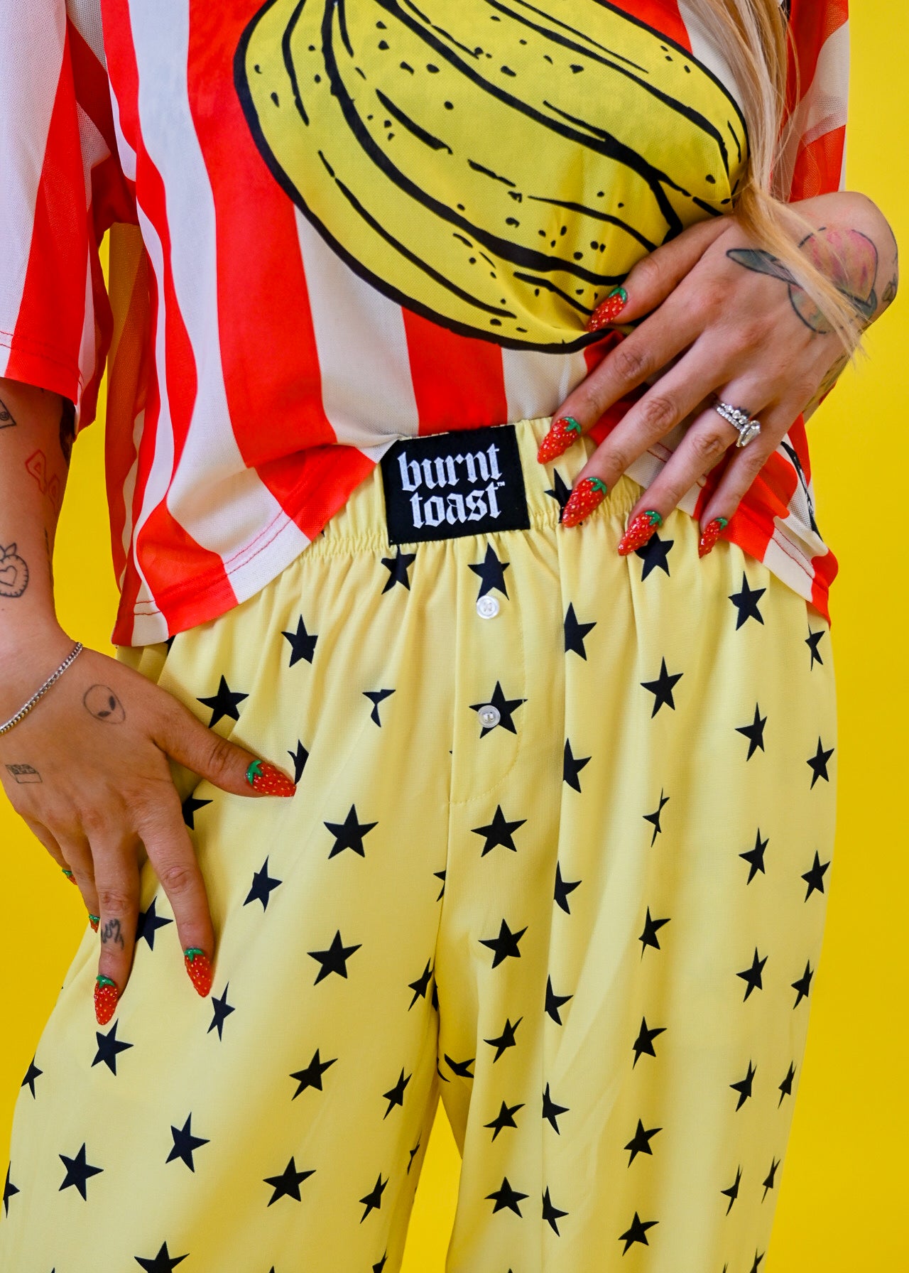 A person wearing yellow pants with a black star pattern and a red-and-white striped shirt with a banana graphic, standing against a bright yellow background. A black waistband label reads 'burnt toast'