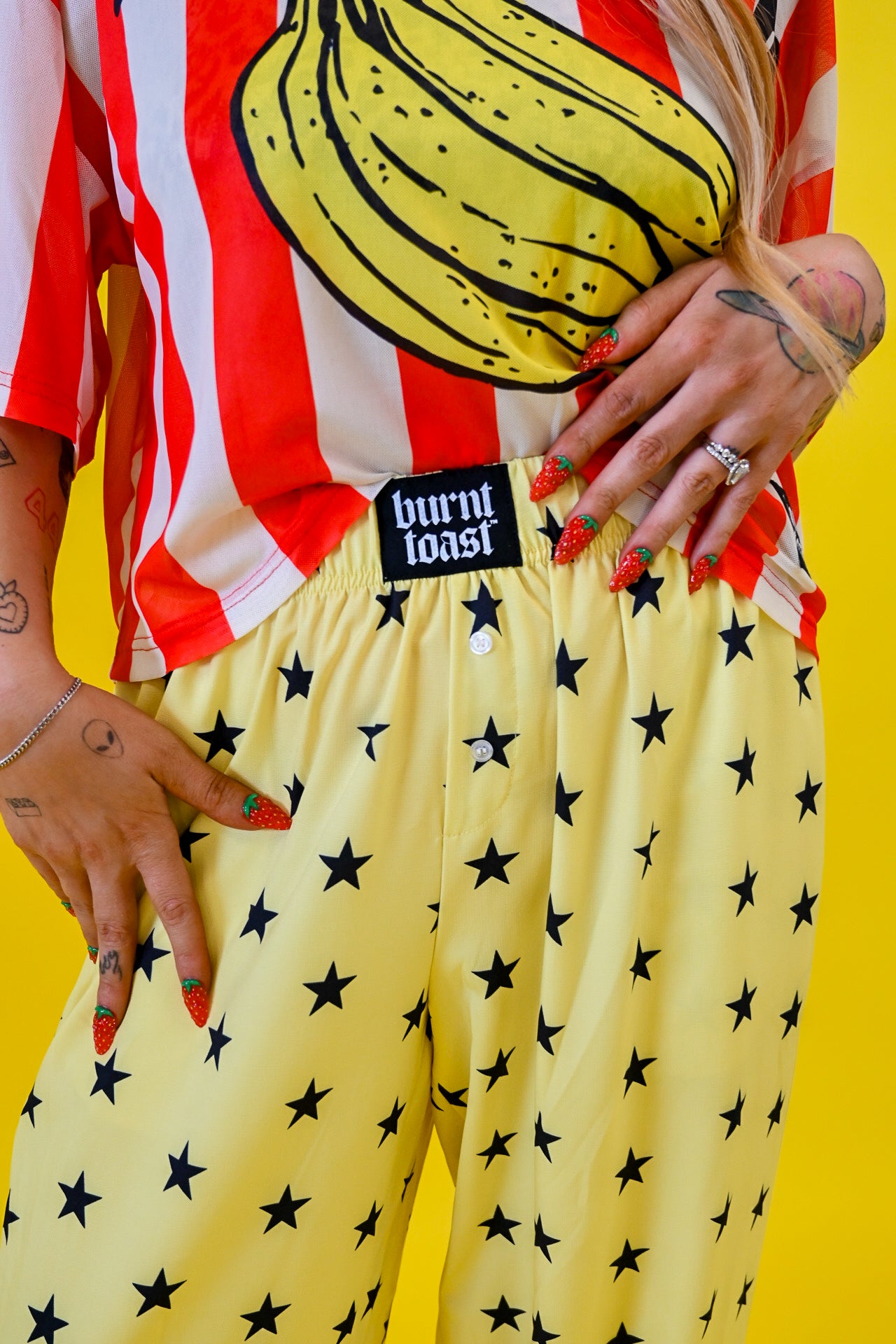 A person wearing yellow pants with a black star pattern and a red-and-white striped shirt with a banana graphic, standing against a bright yellow background. A black waistband label reads 'burnt toast'