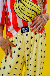 A person wearing yellow pants with a black star pattern and a red-and-white striped shirt with a banana graphic, standing against a bright yellow background. A black waistband label reads 'burnt toast'