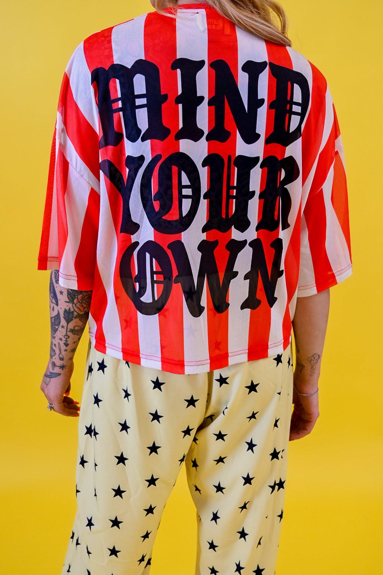 A person shown from the back wearing a red and white vertically striped short-sleeve jersey with large black text reading 'MIND YOUR OWN' across the back, paired with light cream pants covered in black stars, against a solid yellow background