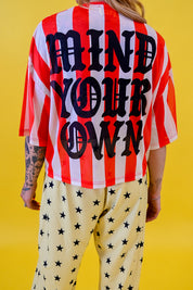 A person shown from the back wearing a red and white vertically striped short-sleeve jersey with large black text reading 'MIND YOUR OWN' across the back, paired with light cream pants covered in black stars, against a solid yellow background