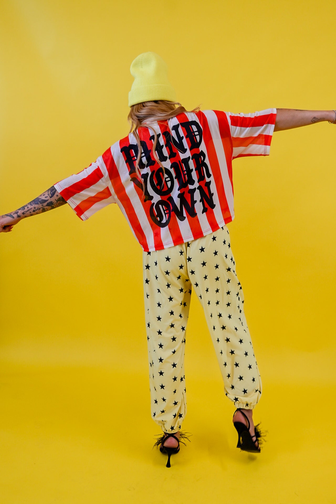 A model from behind wearing a red and white striped jersey with 'MIND YOUR OWN' printed in black, yellow pants with black star prints, a yellow beanie, and black high heels against a solid yellow background