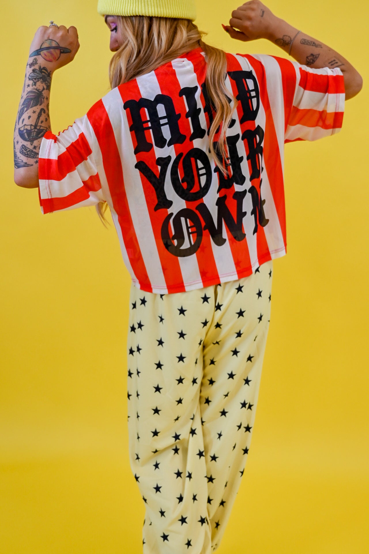 A person shown from the back wearing a red and white striped short-sleeve jersey with large black text reading 'MIND YOUR OWN' and cream pants with black star print, posed against a bright yellow studio background
