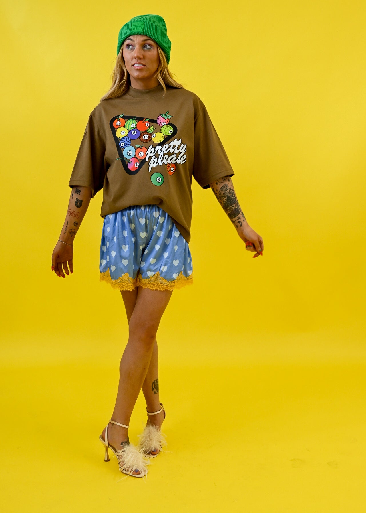 A woman modeling a brown graphic t-shirt with a colorful bowling pin design and the words 'pretty pins', paired with blue patterned shorts, a green beanie, and white high heels, set against a solid yellow background