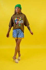 A woman modeling a brown graphic t-shirt with a colorful bowling pin design and the words 'pretty pins', paired with blue patterned shorts, a green beanie, and white high heels, set against a solid yellow background