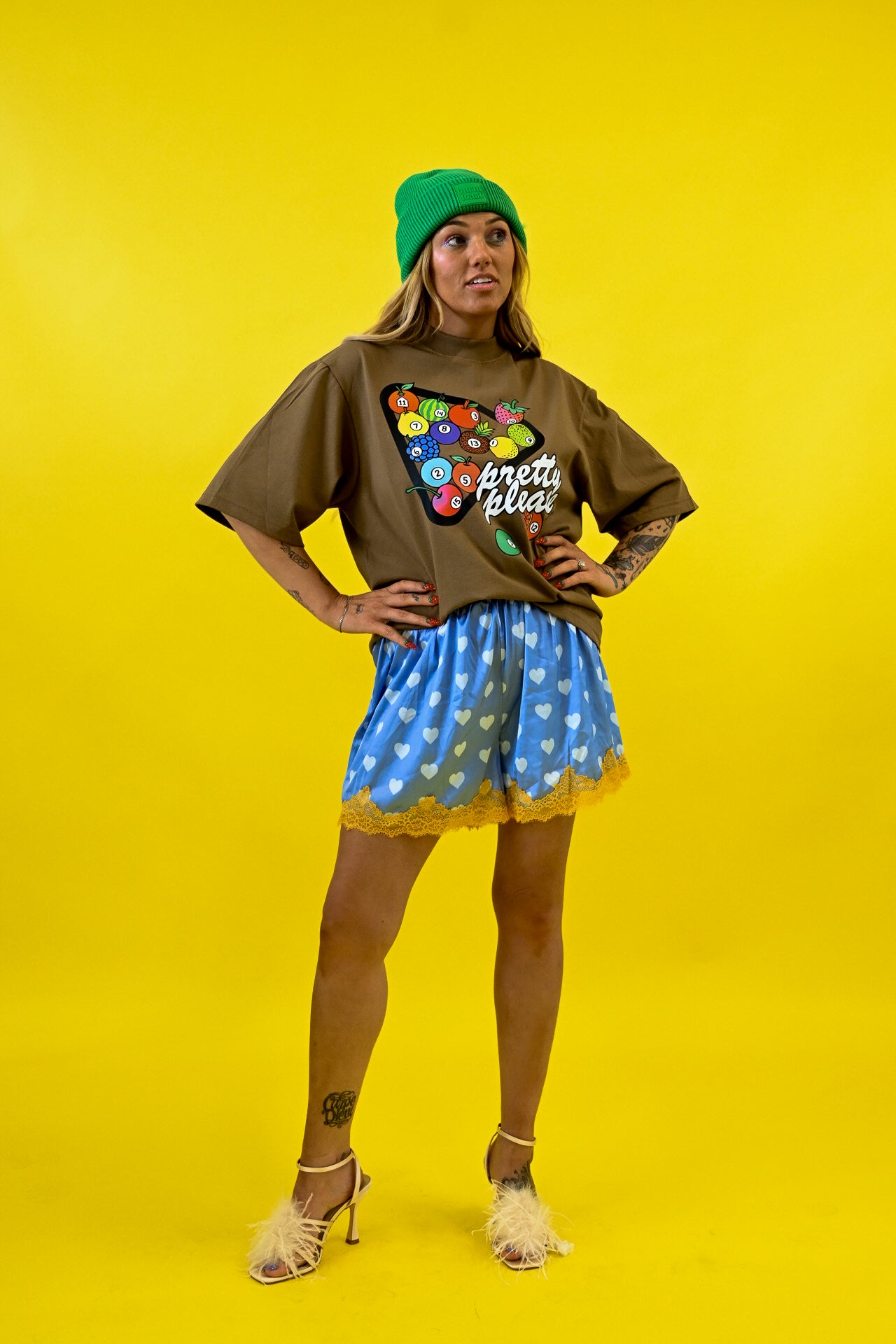 A model posing in a brown oversized t-shirt with a colorful retro graphic and the text 'pretty please', paired with blue shorts featuring a white heart pattern and a yellow ruffled hem, a green beanie, and beige high heels, set against a solid yellow background