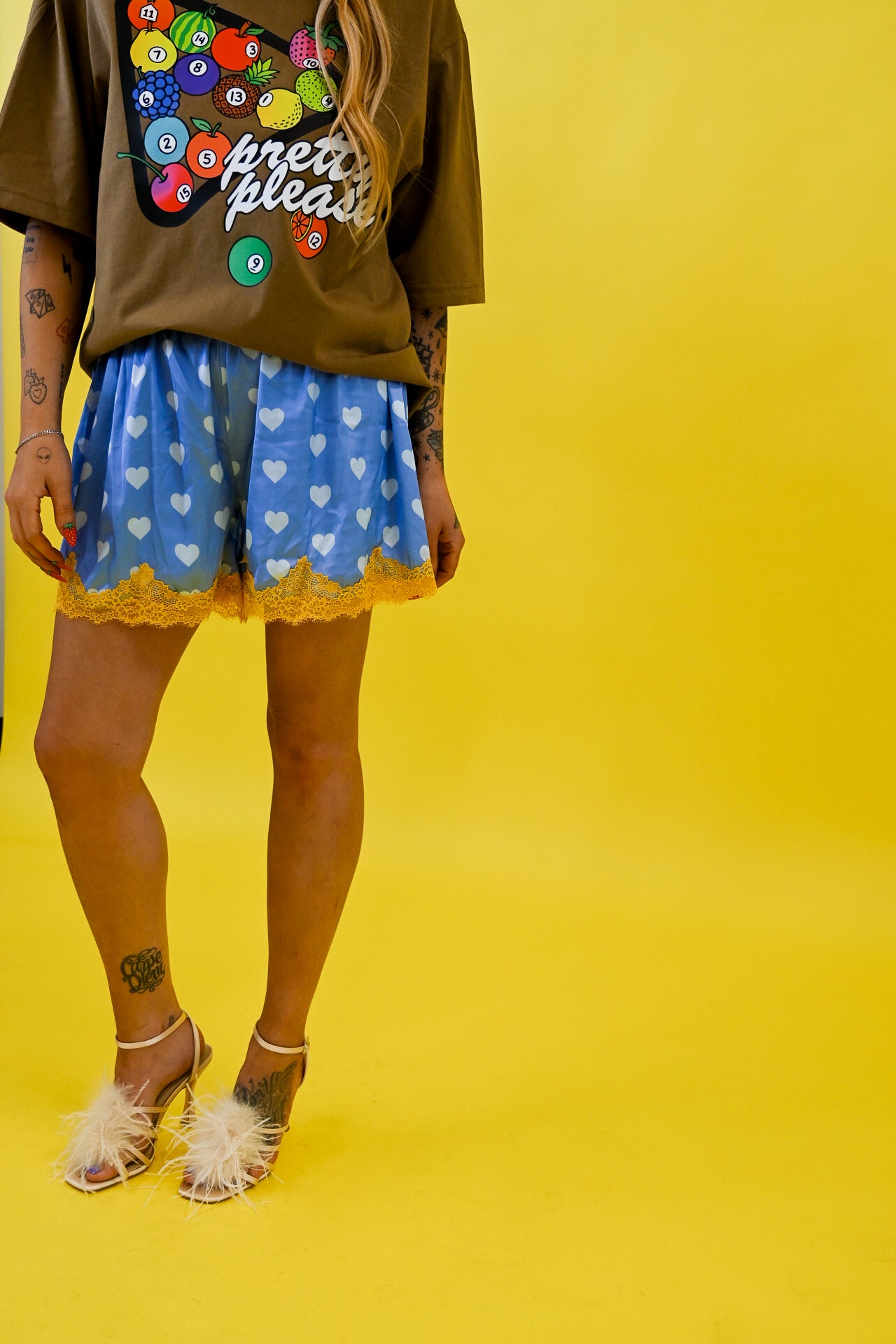 A model wearing blue shorts with a white heart pattern and yellow lace trim, paired with a brown graphic t-shirt and white feather sandals, set against a solid yellow background