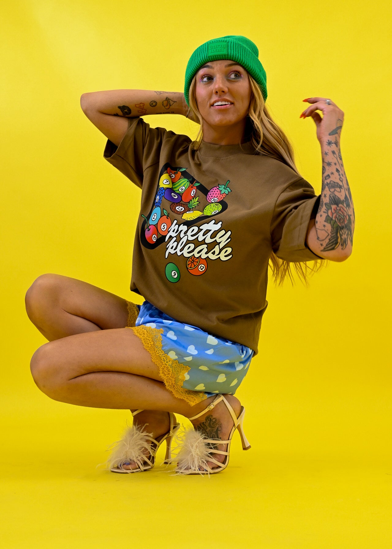 A woman crouching against a bright yellow studio background, wearing a brown graphic T-shirt with colorful pool ball illustrations and the words 'pretty please', a green beanie, blue patterned shorts, and beige high heels 