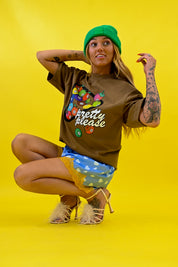 A woman crouching against a bright yellow studio background, wearing a brown graphic T-shirt with colorful pool ball illustrations and the words 'pretty please', a green beanie, blue patterned shorts, and beige high heels 