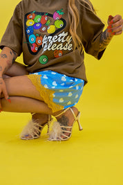 A model wearing a brown graphic t-shirt with a colorful fruit bowl design and the words 'pretty please', paired with a blue and white patterned skirt and beige strappy heels, posed against a solid bright yellow background