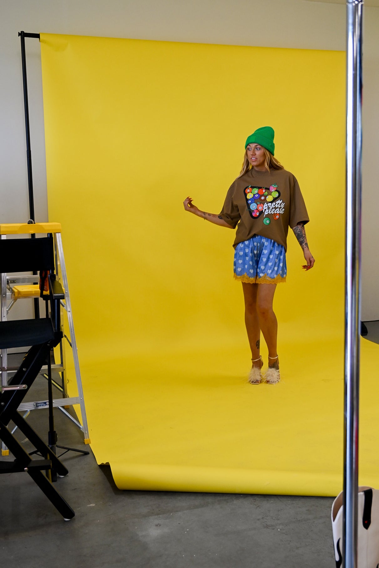 A model stands in front of a bright yellow photography backdrop wearing a brown graphic t-shirt, blue patterned shorts, and a green beanie. Studio equipment, including a ladder and support pole, is visible on the left and right edges