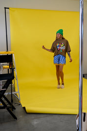 A model stands in front of a bright yellow photography backdrop wearing a brown graphic t-shirt, blue patterned shorts, and a green beanie. Studio equipment, including a ladder and support pole, is visible on the left and right edges