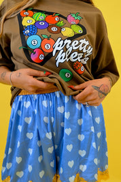 A person wearing a brown graphic T-shirt with colorful fruit illustrations and the words 'pretty please' in white script, paired with a blue lace shorties patterned with white hearts, set against a solid yellow background