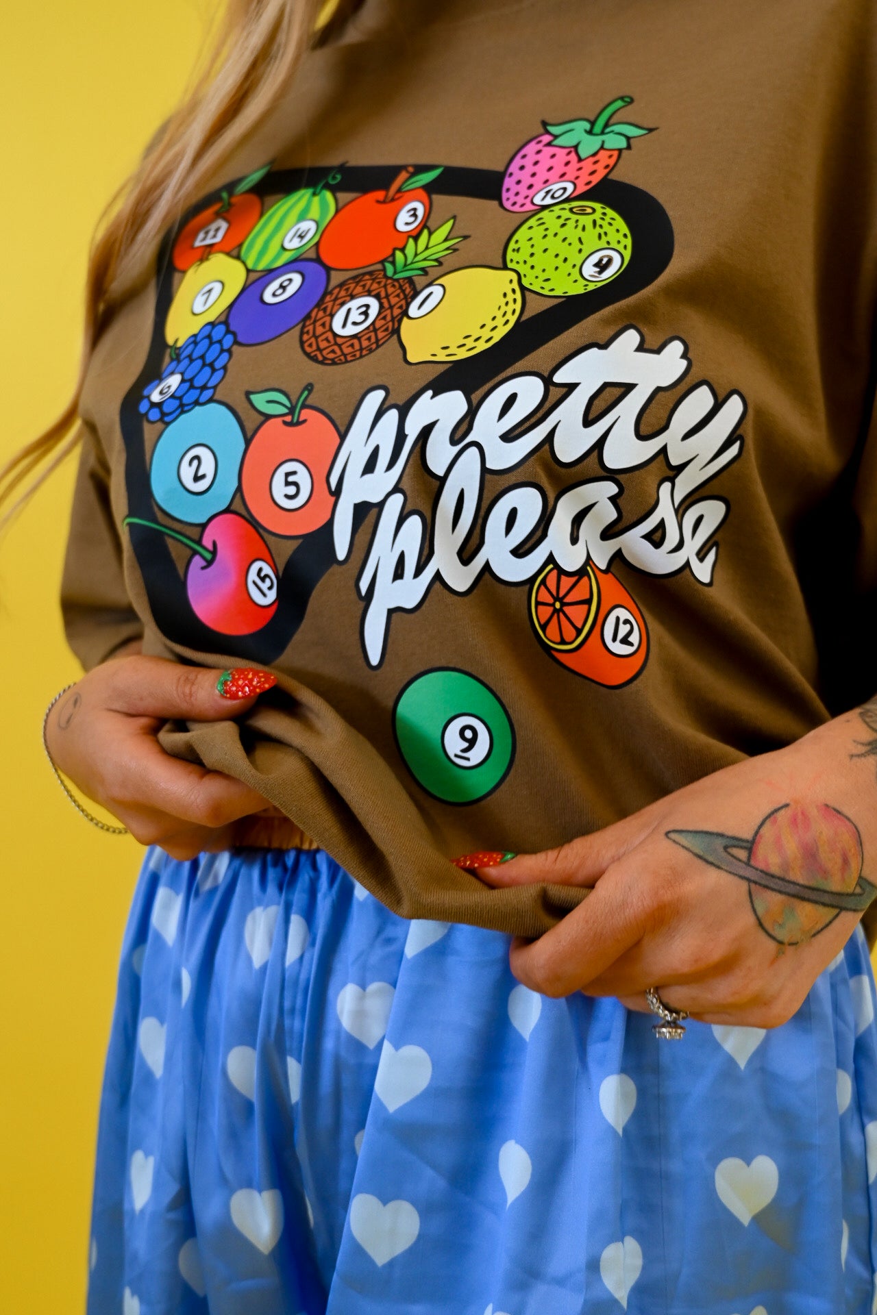 A person wearing a brown graphic T-shirt with a colorful fruit bowl design and the words 'pretty please' in white script, paired with a blue skirt with white hearts, against a yellow background
