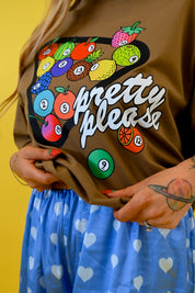 A person wearing a brown graphic T-shirt with a colorful fruit bowl design and the words 'pretty please' in white script, paired with a blue skirt with white hearts, against a yellow background