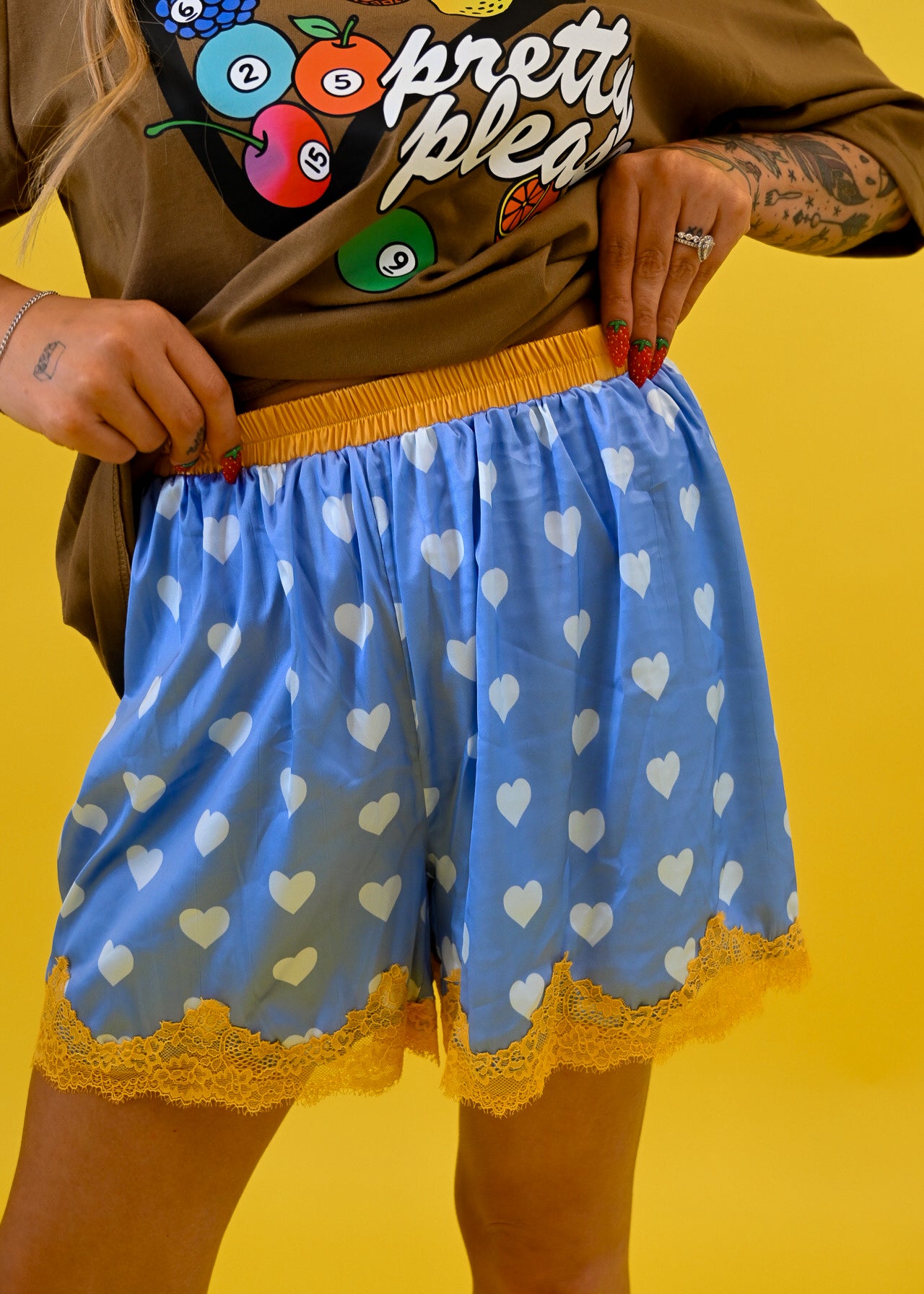 A person wearing light blue shorts with a white heart pattern, yellow elastic waistband, and yellow lace hem, paired with a brown graphic t-shirt, set against a solid yellow background, close up view
