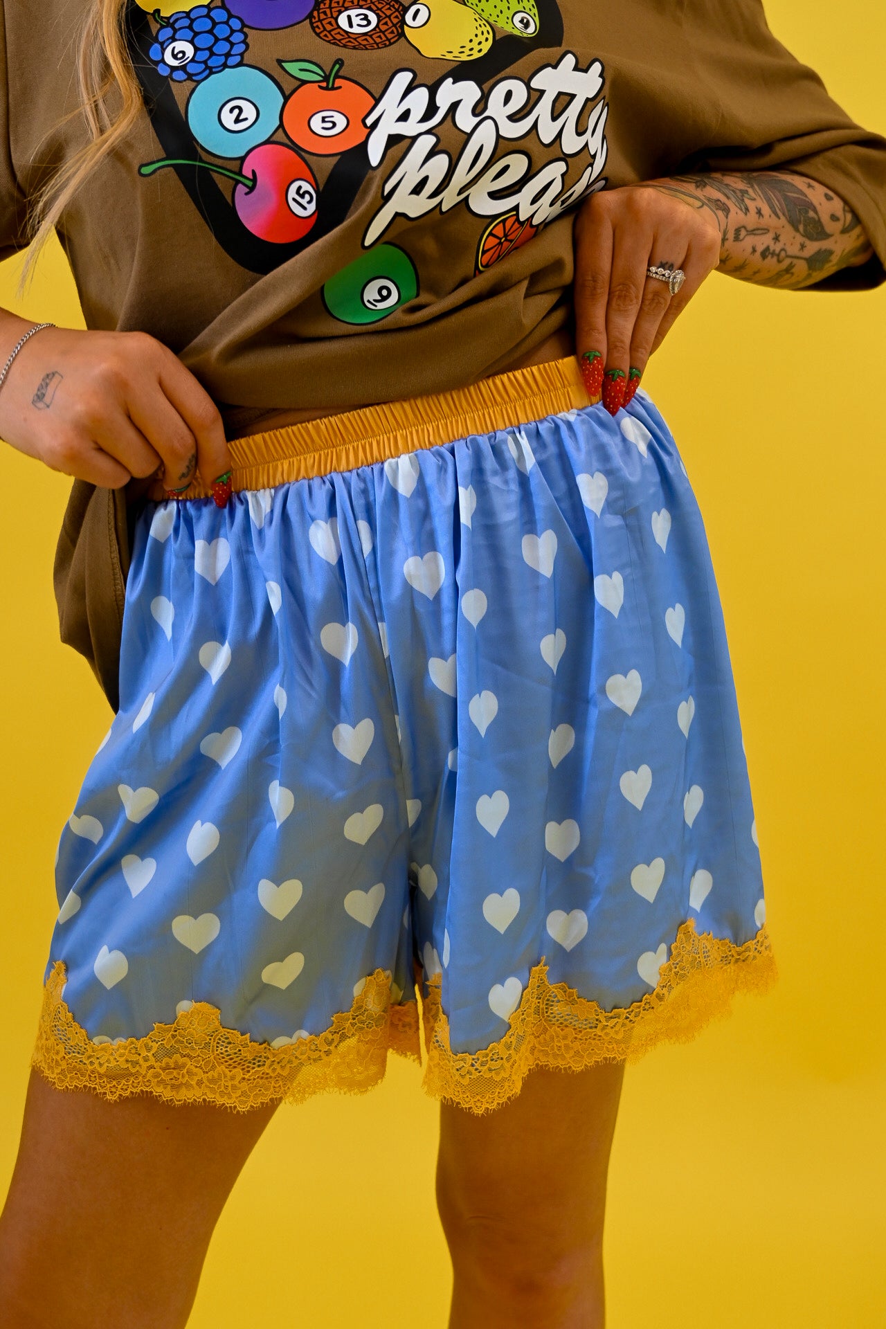 A person wearing light blue shorts with a white heart pattern, yellow elastic waistband, and yellow lace hem, paired with a brown graphic t-shirt, set against a solid yellow background, close up view