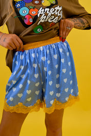 A person wearing light blue shorts with a white heart pattern, yellow elastic waistband, and yellow lace hem, paired with a brown graphic t-shirt, set against a solid yellow background, close up view