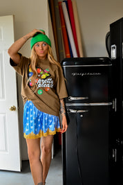 A woman modeling a brown graphic t-shirt with colorful candy graphics and the words 'pretty please', paired with a blue and white patterned skirt, a green beanie, and standing next to a black retro refrigerator