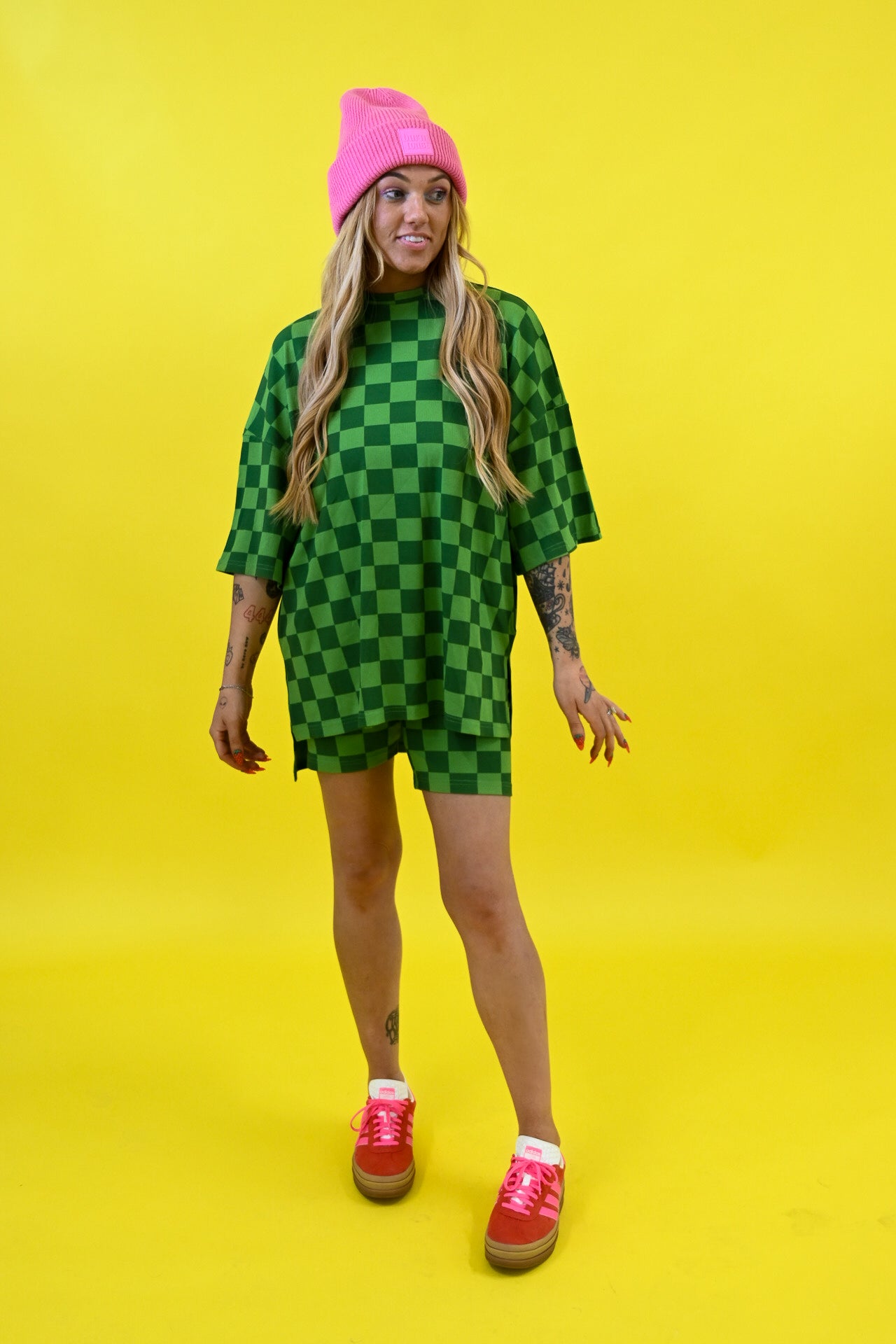 A woman modeling an oversized green checkered t-shirt dress with a pink beanie and pink sneakers, standing against a solid bright yellow background