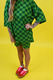 A person wearing an oversized green and black checkered short-sleeve top and matching shorts, paired with bright pink sneakers, standing against a solid yellow background, close up view