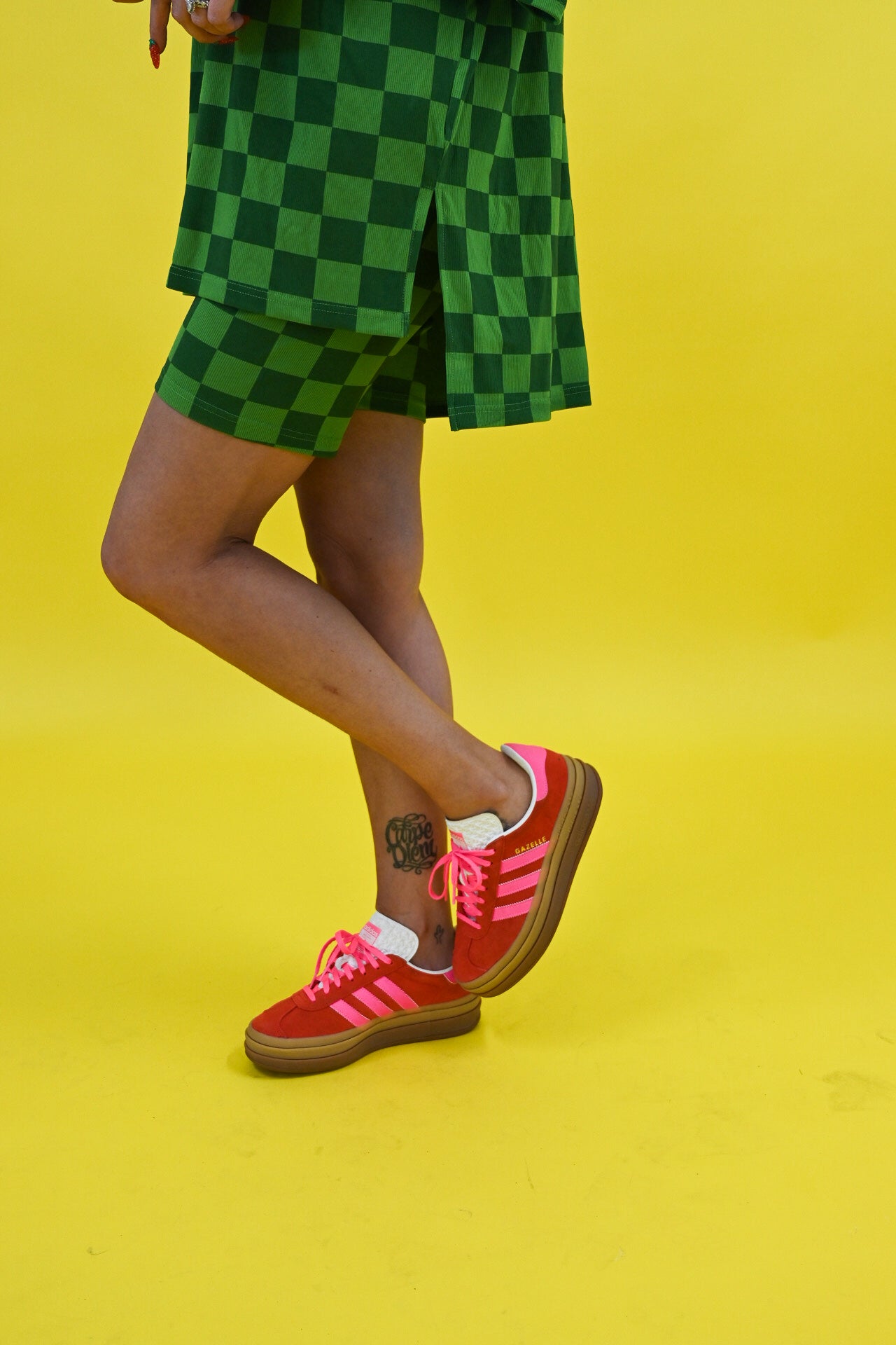 A person wearing green checkered shorts and a matching top, paired with red Adidas sneakers with pink stripes, standing against a bright yellow background, close up side view