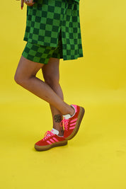 A person wearing green checkered shorts and a matching top, paired with red Adidas sneakers with pink stripes, standing against a bright yellow background, close up side view