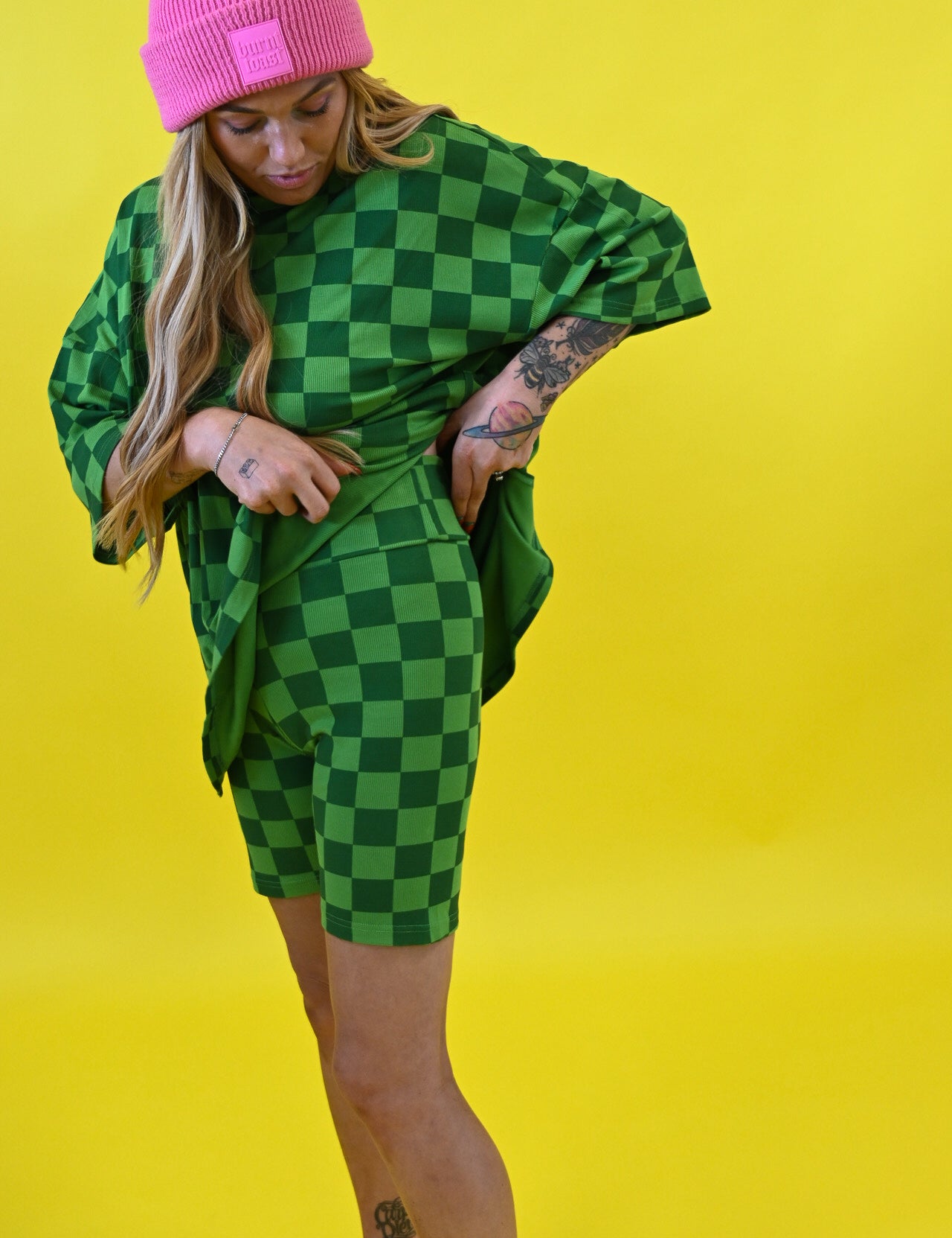 A woman posing in a green and black checkered oversized t-shirt and matching shorts, paired with a bright pink beanie and pink sneakers, set against a solid yellow background side view