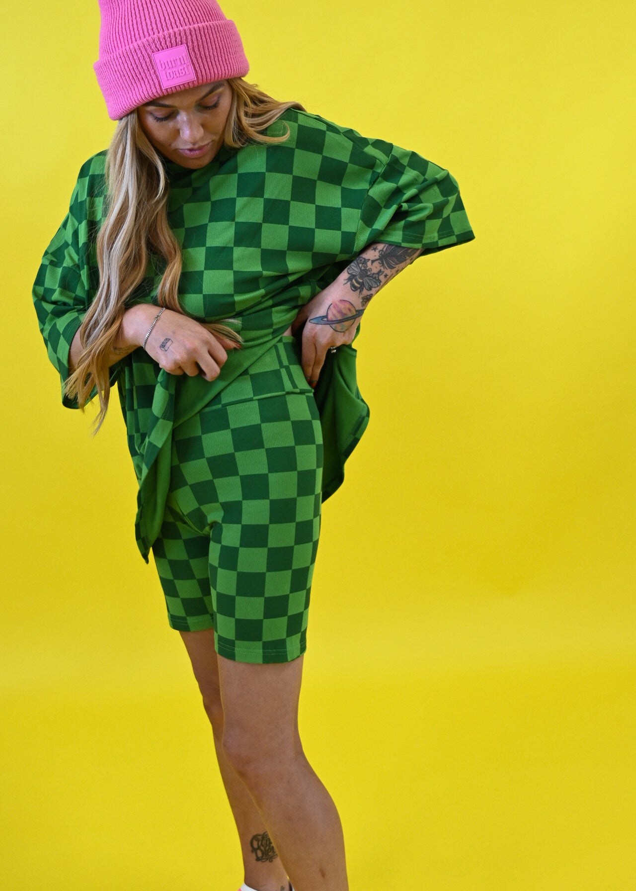A woman posing in a green and black checkered oversized t-shirt and matching shorts, paired with a bright pink beanie and pink sneakers, set against a solid yellow background side view