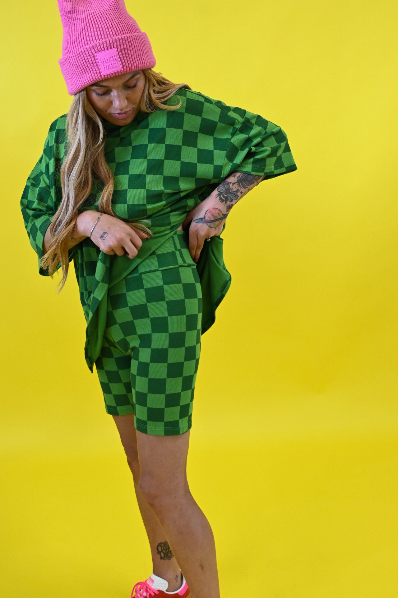 A woman posing in a green and black checkered oversized t-shirt and matching shorts, paired with a bright pink beanie and pink sneakers, set against a solid yellow background side view