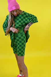 A woman posing in a green and black checkered oversized t-shirt and matching shorts, paired with a bright pink beanie and pink sneakers, set against a solid yellow background side view