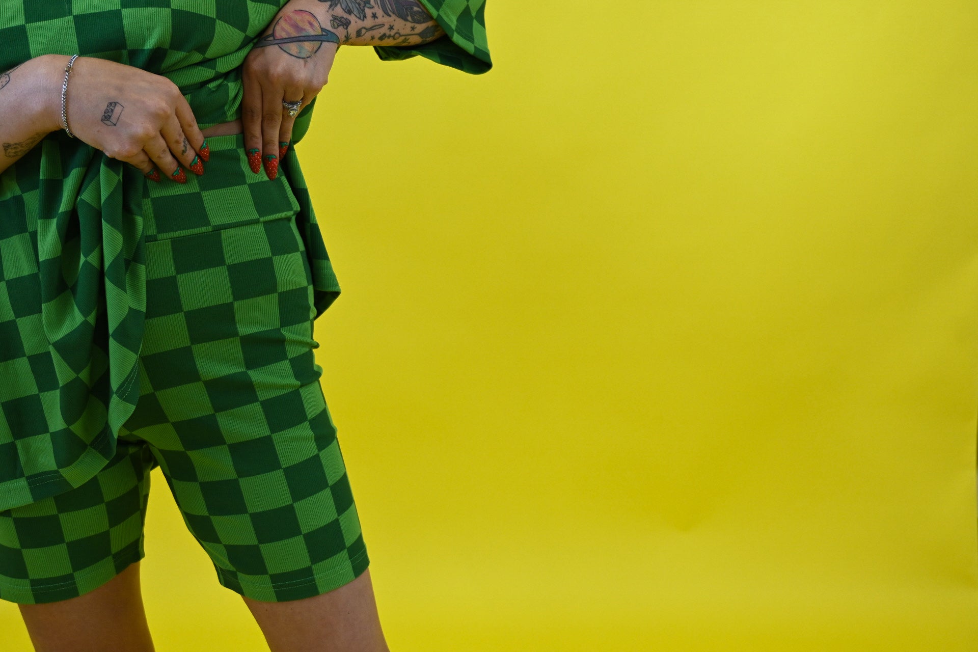 A person wearing a green and dark green checkered short-sleeve top and matching shorts, standing against a solid bright yellow background. The person is cropped from the waist down, focusing on waistband