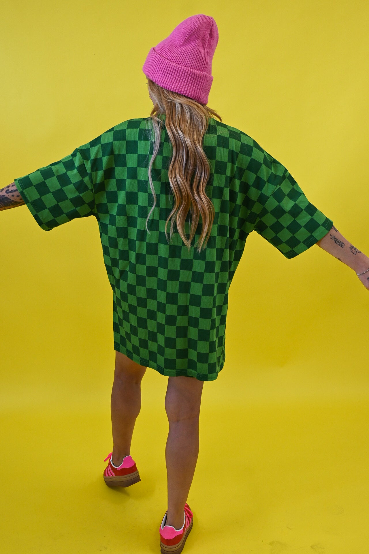 A person shown from behind wearing an oversized green and black checkered t-shirt dress, a pink beanie, and pink sneakers against a solid yellow background
