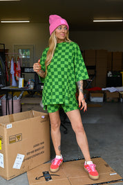 A woman stands in a garage-like storage area wearing an oversized green-and-yellow checkered tunic top and matching checkered shorts, with a bright pink beanie and pink sneakers. Cardboard boxes are visible around her