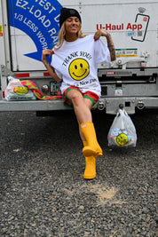 A woman sits on the back of a U-Haul truck wearing a white graphic T-shirt with a yellow smiley face and the words 'THANK YOU! You & Nice Day', red patterned shorts, and bright yellow rain boots. She holds plastic bags, with one bag featuring a matching yellow smiley face design