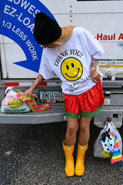 A person standing in front of a white truck tailgate wearing a white graphic T-shirt with a large yellow smiley face and the words 'THANK YOU!', a black beanie, a red strawberry-print skirt, and bright yellow rain boots. Bags are hanging near the person and truck