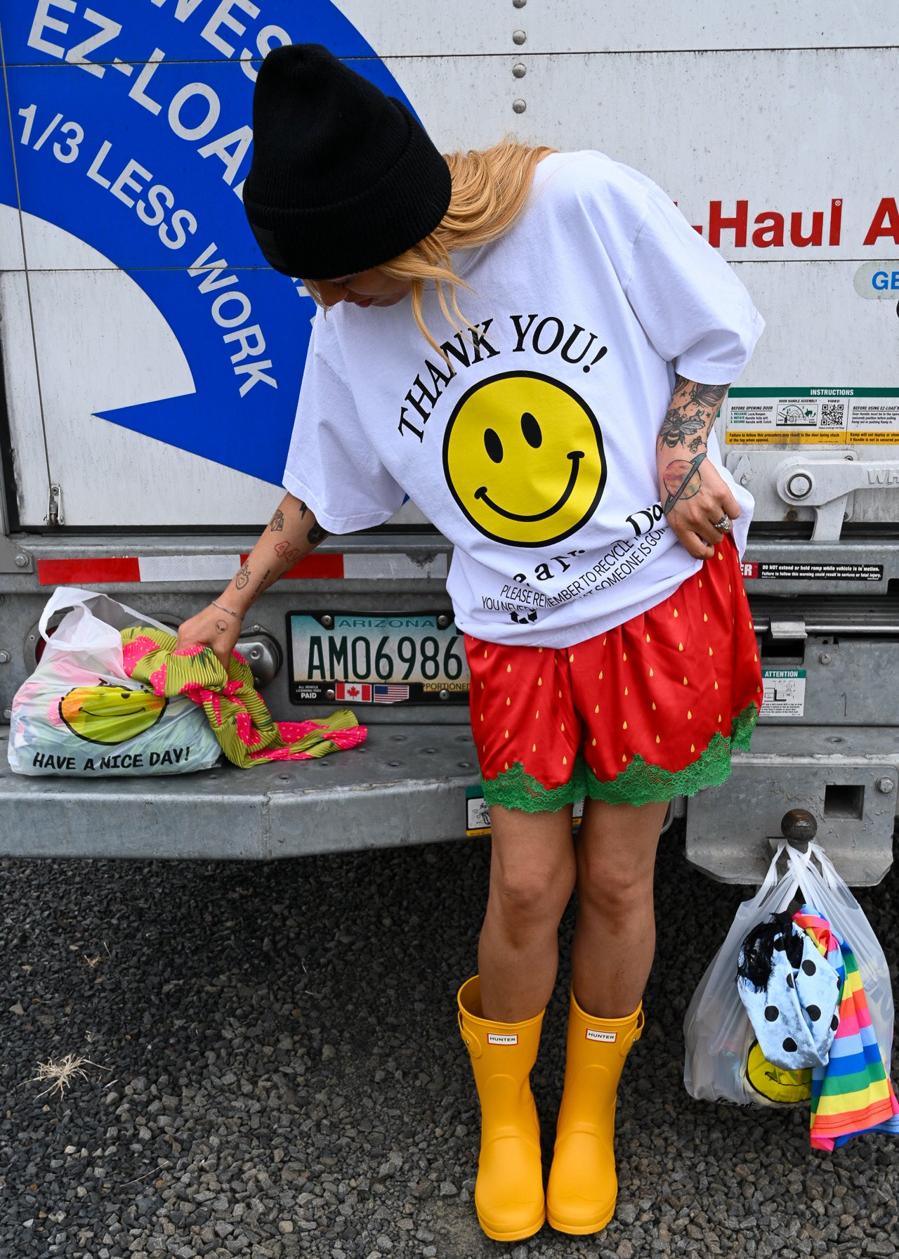 A person standing in front of a white truck tailgate wearing a white graphic T-shirt with a large yellow smiley face and the words 'THANK YOU!', a black beanie, a red strawberry-print skirt, and bright yellow rain boots. Bags are hanging near the person and truck