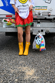 A person standing outdoors in front of a truck, wearing a white graphic T-shirt, a red and green patterned skirt, and bright yellow rain boots. A plastic bag with colorful clothing hangs from the hitch beside them on gravel