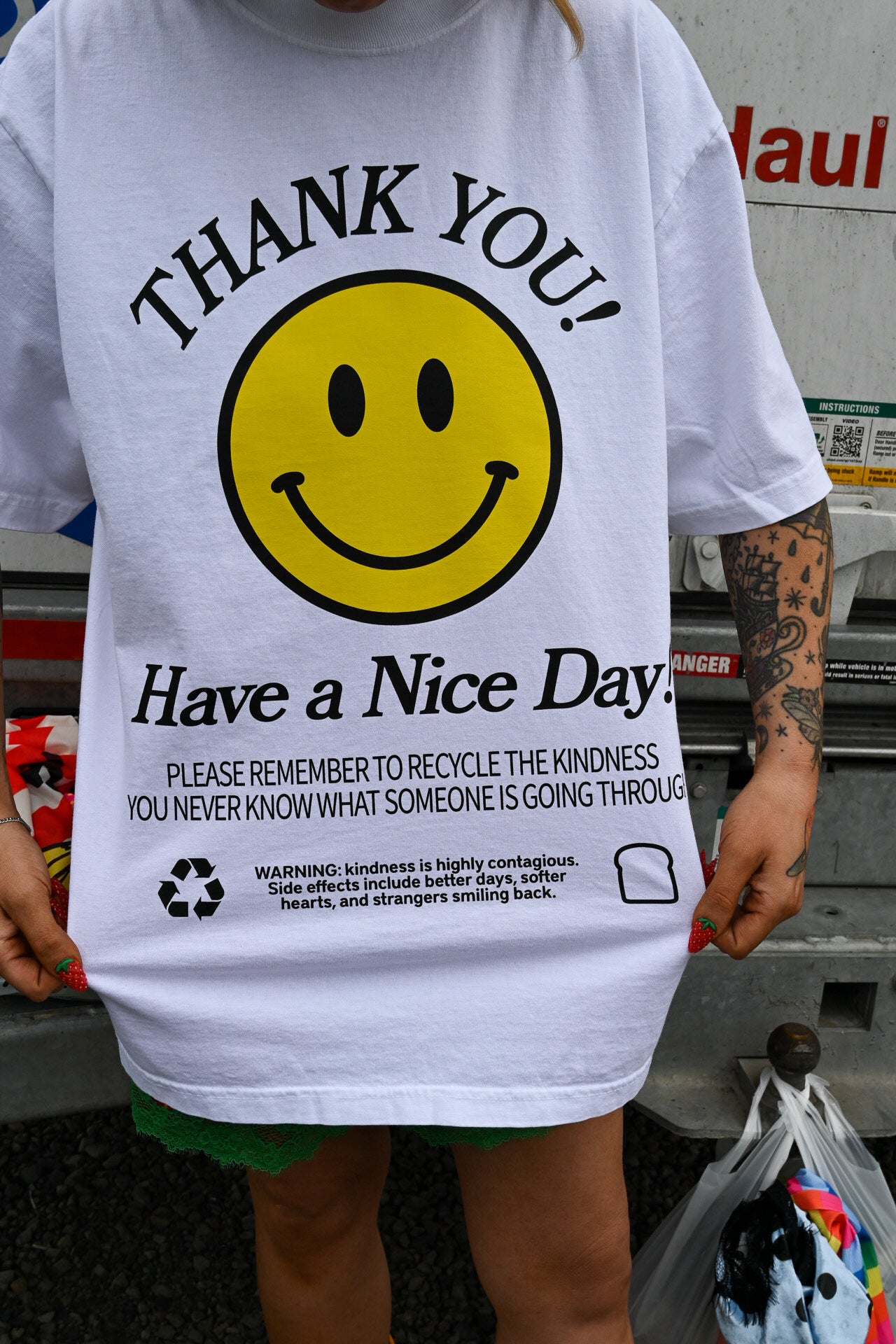 A person wearing a white graphic T-shirt with a large yellow smiley face and black text reading 'THANK YOU! Have a Nice Day!' and additional recycling-themed text, photographed outdoors near a truck