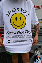 A person wearing a white graphic T-shirt with a large yellow smiley face and black text reading 'THANK YOU! Have a Nice Day!' and additional recycling-themed text, photographed outdoors near a truck