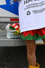 A person wearing a white graphic T-shirt with recycling text and a red strawberry-patterned skirt with green lace trim, standing in front of a vehicle