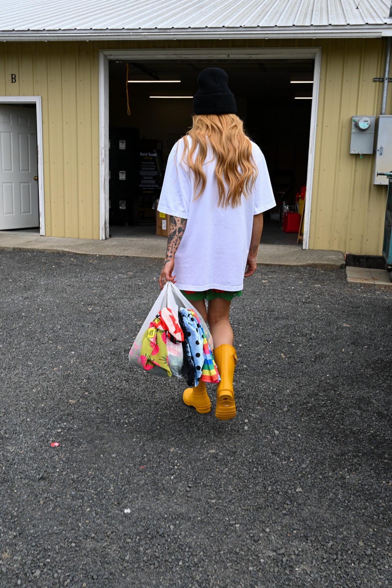 A person seen from behind walking toward an open garage door, wearing a white oversized T-shirt, black beanie, green shorts, bright yellow rain boots, and holding a clear plastic bag filled with colorful items