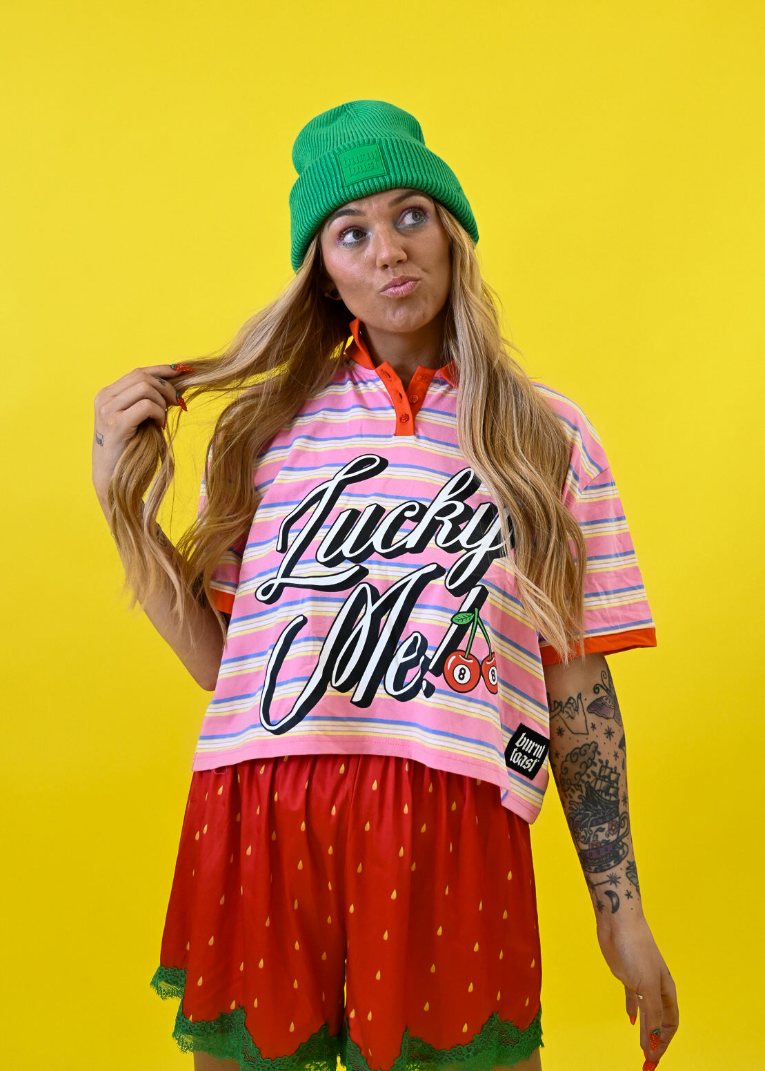 A woman posing in a vibrant studio setting against a solid yellow background, wearing a green beanie, a pink and white striped t-shirt with 'Lucky Me!' text, and red strawberry-patterned shorts
